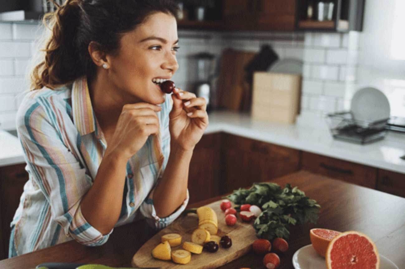Mulher sentada numa bancada em sua cozinha, comendo uma fruta