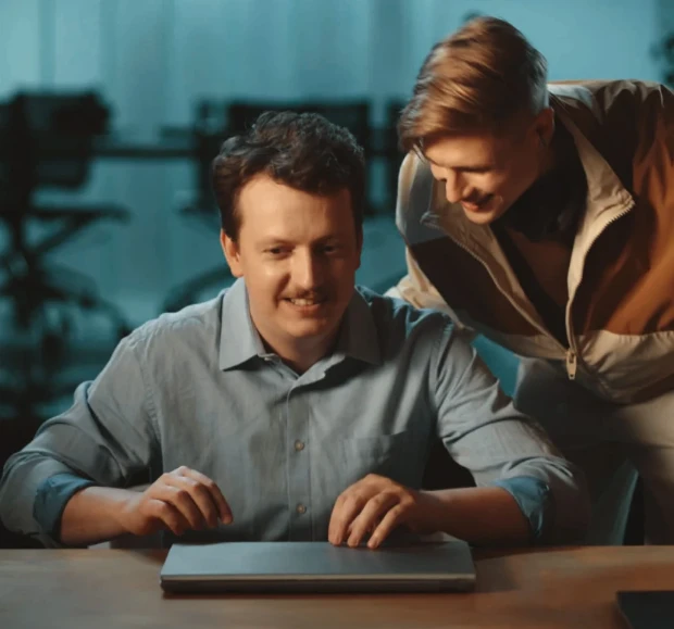 Dois colegas de trabalho sorrindo em um escritório. Um deles está com a mão no laptop fechado.