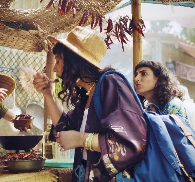 Duas mulheres provando comida de rua sob um mercado com pimentas secas penduradas.