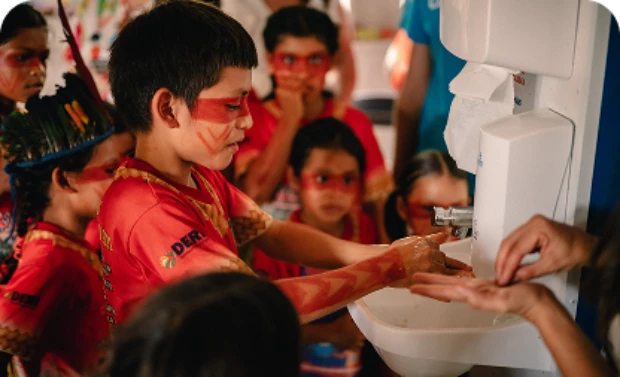 Crianças com pintura facial lavam as mãos em uma pia com orientação.