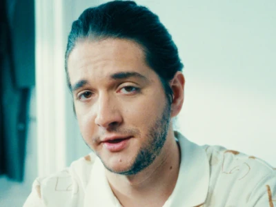 Homem de cabelos e barba escuros, vestindo camisa de colarinho de cor clara, dentro de casa.