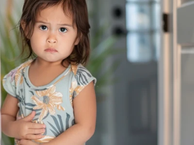 Criança pequena em uma camisa floral com aparência triste, segurando a barriga dentro de casa.