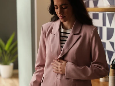 Mulher em um blazer rosa segurando a barriga, em pé em um ambiente fechado.
