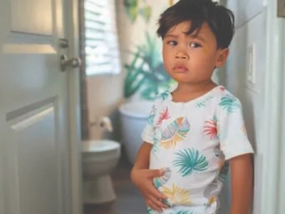 Criança segurando o estômago, em pé perto da porta do banheiro, parecendo preocupada.