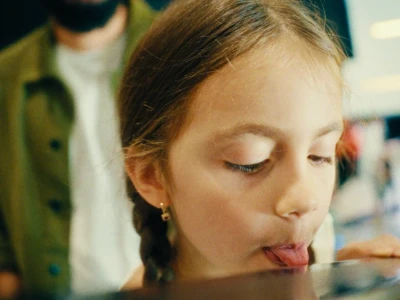 Uma garota lambe uma superfície refletora com um adulto ao fundo.