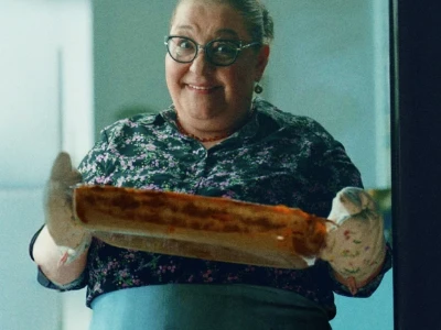 Pessoa sorridente segurando um prato assado com luvas de forno em uma cozinha.