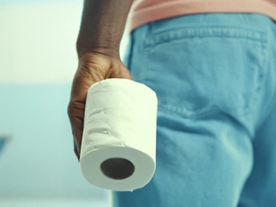 Pessoa sentada em um vaso sanitário segurando um rolo de papel higiênico em um banheiro.