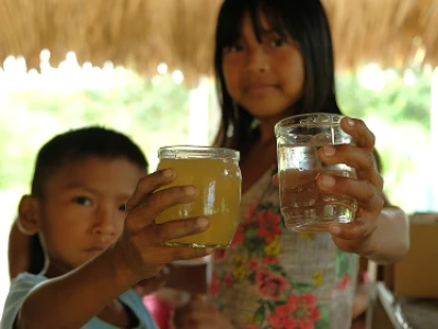 Criança segurando dois copos, um com uma bebida amarela e outro com água, sob um telhado de palha.