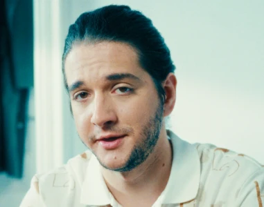 Homem de cabelos e barba escuros, vestindo camisa de colarinho de cor clara, dentro de casa.