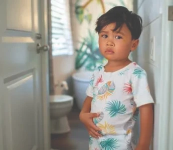 Criança segurando o estômago, em pé perto da porta do banheiro, parecendo preocupada.