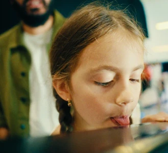 Uma garota lambe uma superfície refletora com um adulto ao fundo.