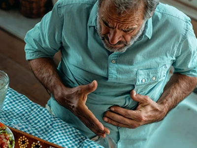 Homem idoso sentado em uma mesa, segurando o estômago, com desconforto.