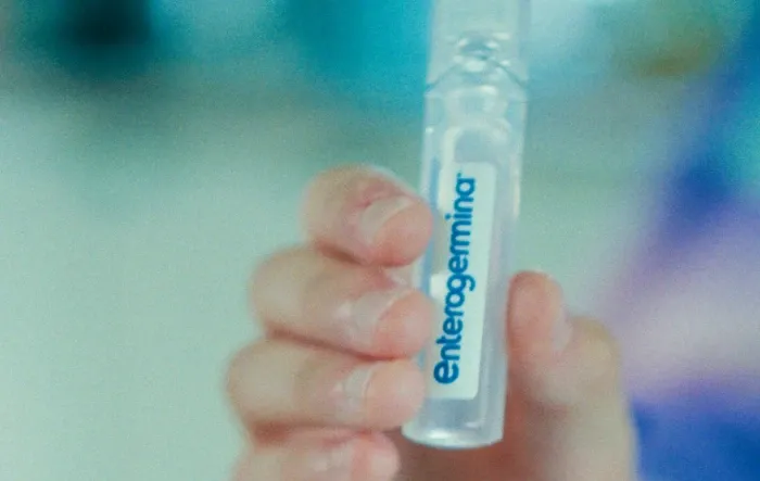 Mão segurando um flaconete de Enterogermina em suspensão oral. Fundo desfocado. 