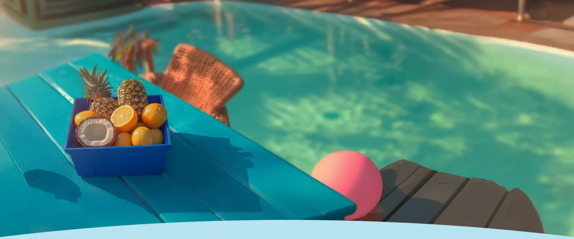Frutas tropicais em uma caixa azul em uma mesa turquesa ao lado de uma piscina com uma bola rosa.