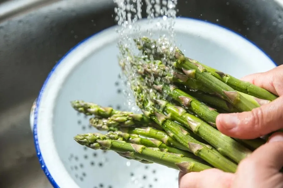 Mãos lavam um maço de espargos verdes sob água corrente em um escorredor branco.