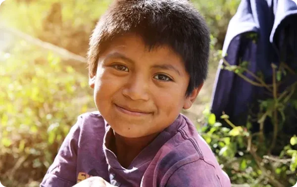 Criança sorridente com uma camisa roxa sentada ao ar livre sob a luz do sol.