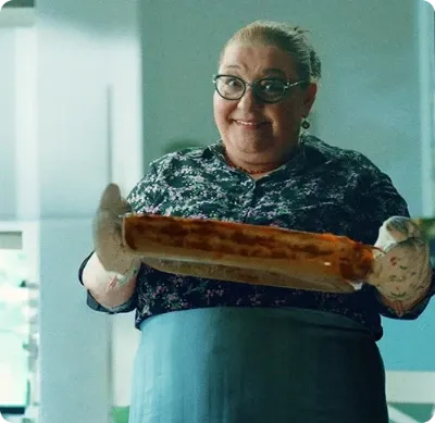 Pessoa sorridente segurando um prato assado com luvas de forno em uma cozinha.