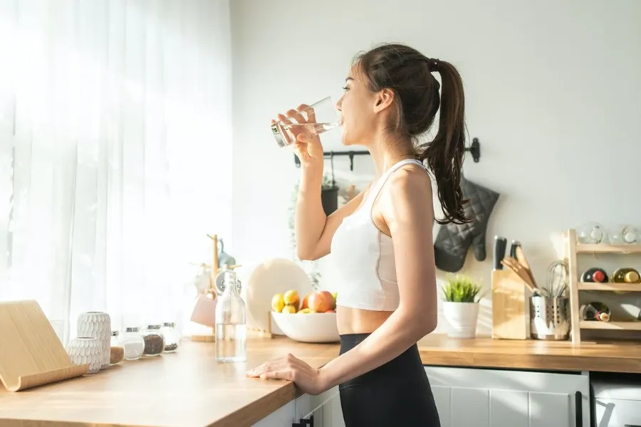 Mulher em traje de ginástica bebendo água em uma cozinha iluminada pelo sol.