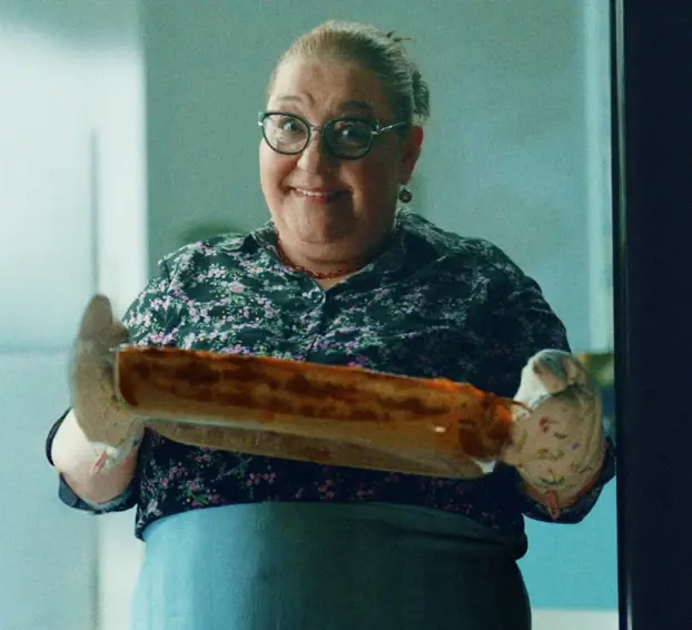 Pessoa sorridente segurando um prato assado com luvas de forno em uma cozinha.