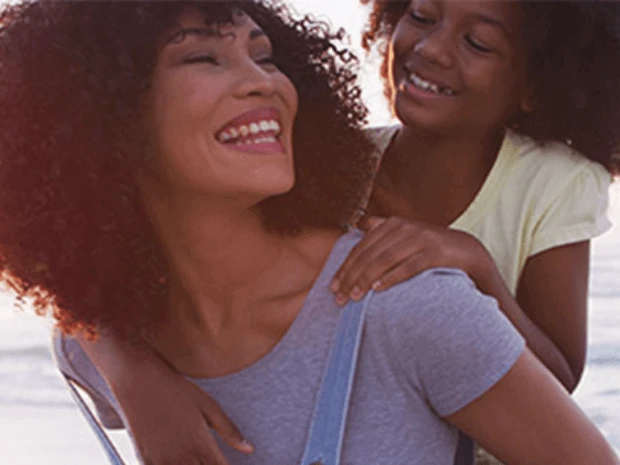 Mãe com a filha nas costas, sorrindo, com o mar ao fundo durante uma viagem