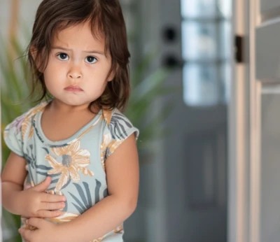 Criança pequena em uma camisa floral com aparência triste, segurando a barriga dentro de casa.