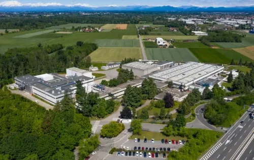 Vista aérea de edifícios industriais cercados por campos verdes e montanhas distantes.
