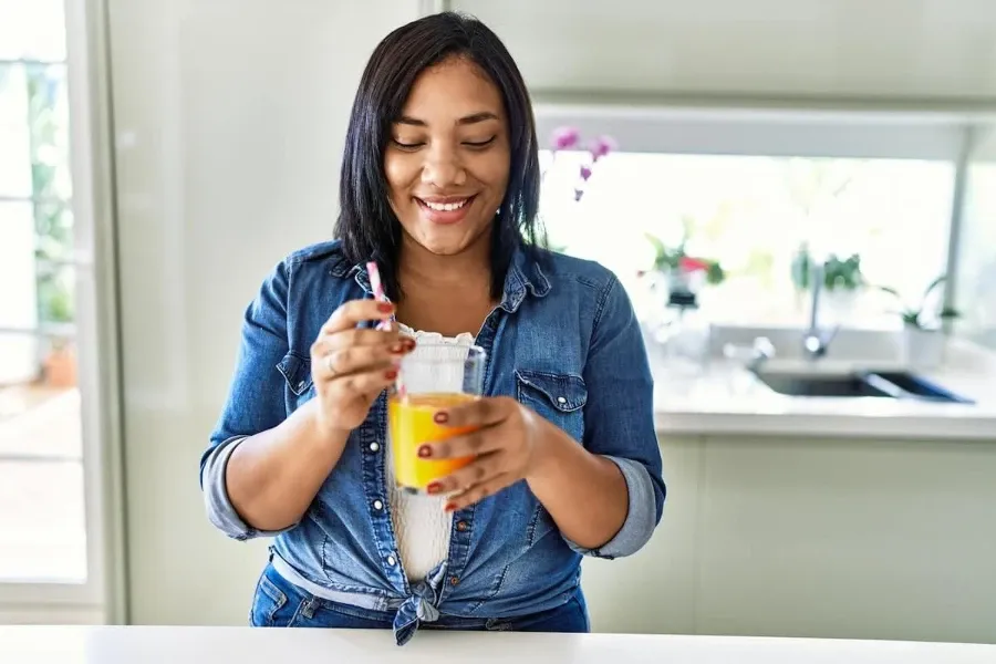 Mulher sorridente de camisa jeans bebendo suco de laranja com canudo em uma cozinha moderna.