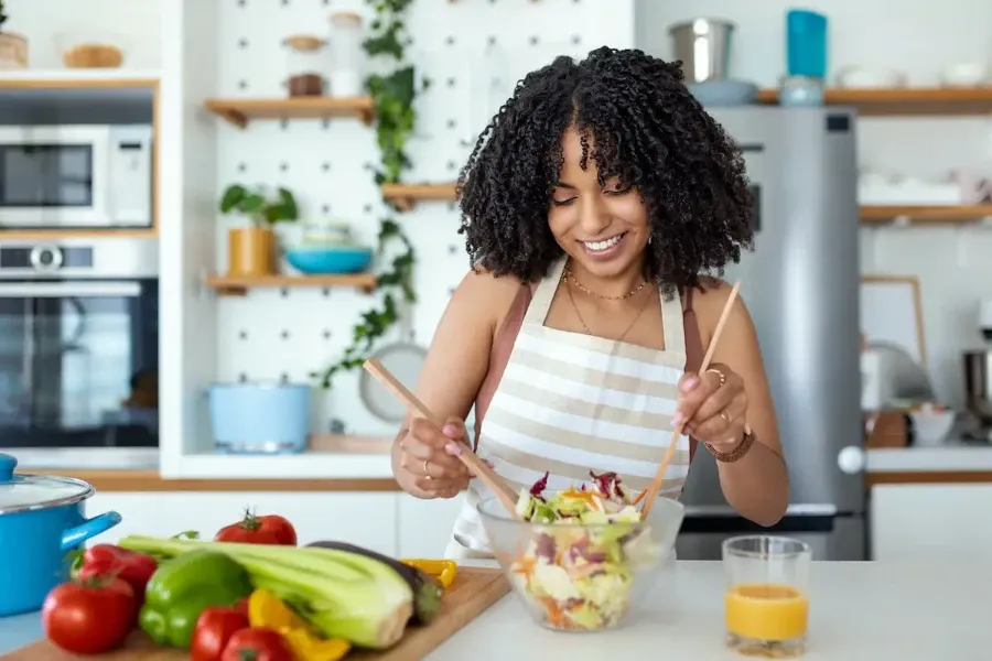 Mulher de avental preparando uma salada em uma cozinha moderna com legumes e suco por perto.