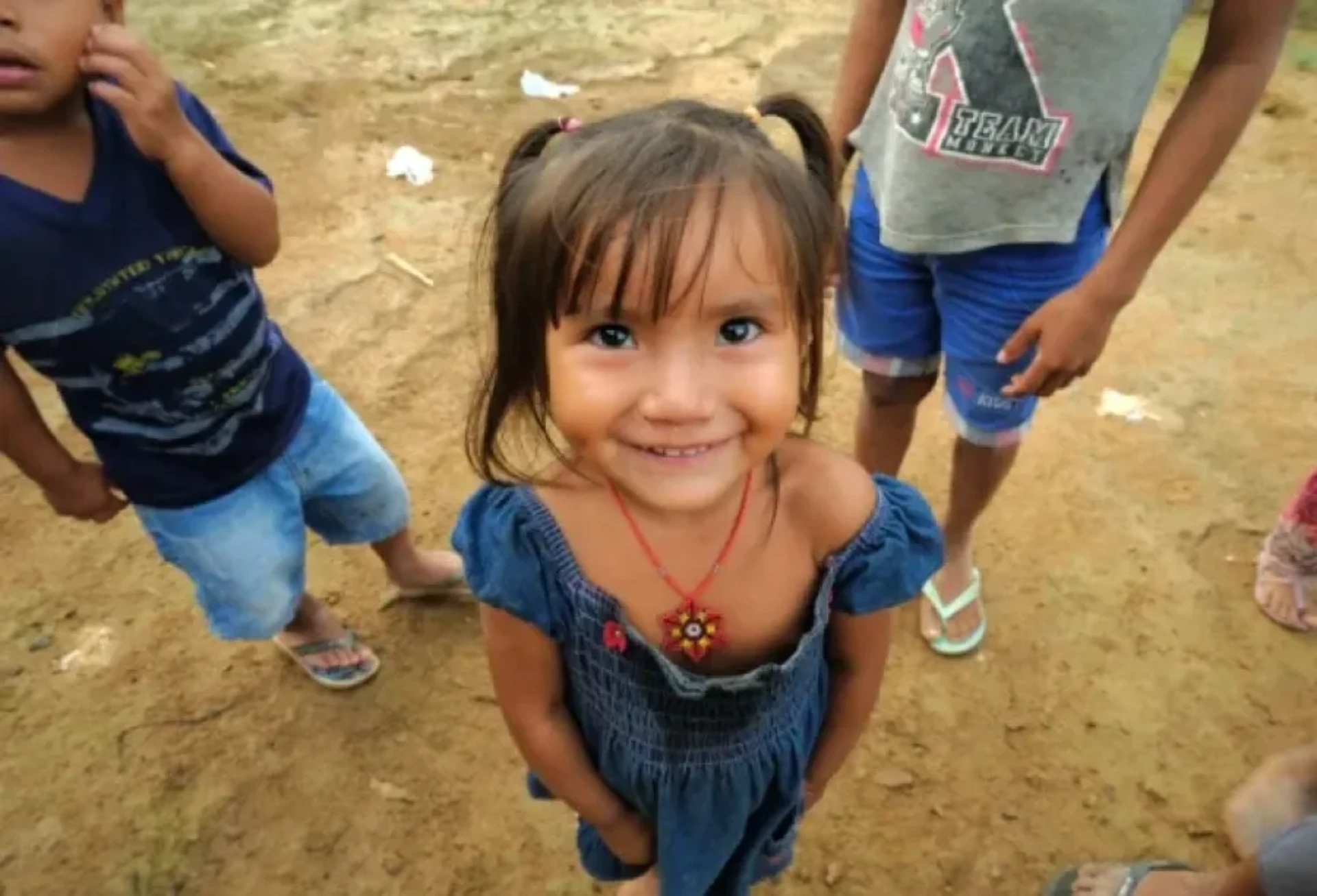 Menina sorridente de vestido azul e colar vermelho, com outras crianças no fundo;