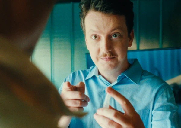 Uma pessoa de camisa azul faz gestos com as mãos em um ambiente fechado.