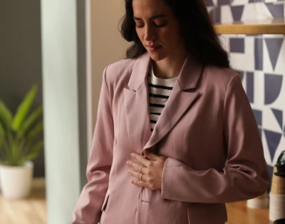 Mulher em um blazer rosa segurando a barriga, em pé em um ambiente fechado.