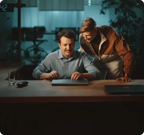 Dois colegas de trabalho sorrindo em um escritório. Um deles está com a mão no laptop fechado.