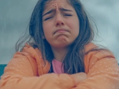 Menina com sobrancelhas franzidas, vestindo uma jaqueta laranja, parada na chuva.