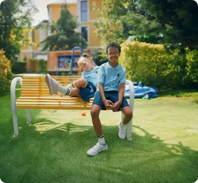 Duas crianças com uniformes azuis combinando, sentadas em um banco de praça.