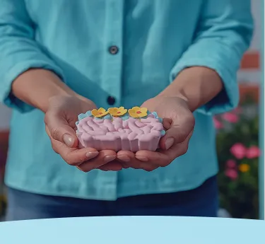 Mãos segurando um modelo de cérebro rosa com decorações de flores amarelas na parte superior.