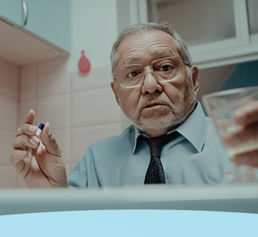 Homem idoso segurando uma cápsula de medicamento e um copo de água em uma cozinha.