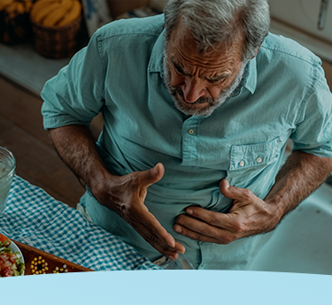 Homem idoso segurando o estômago, parecendo ter desconforto em uma mesa de cozinha.