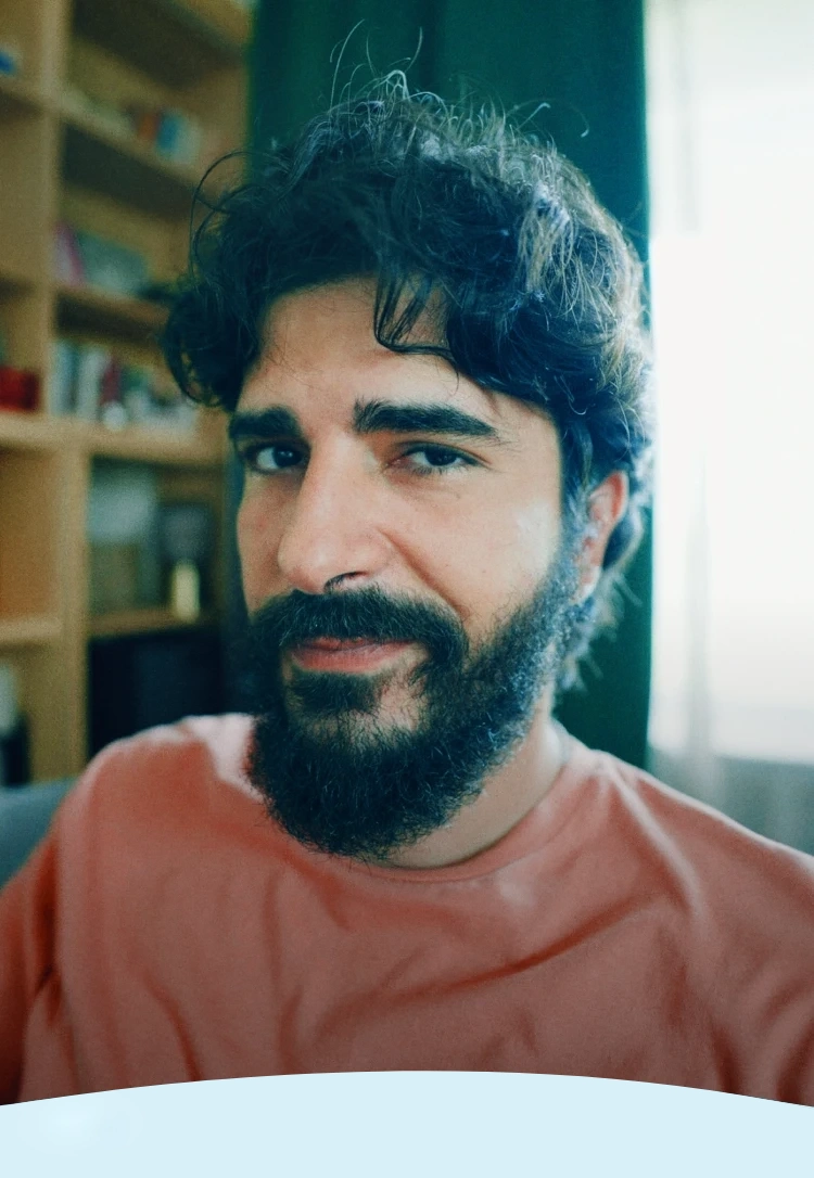 Um homem de cabelos escuros e barba sorrindo levemente dentro de casa.