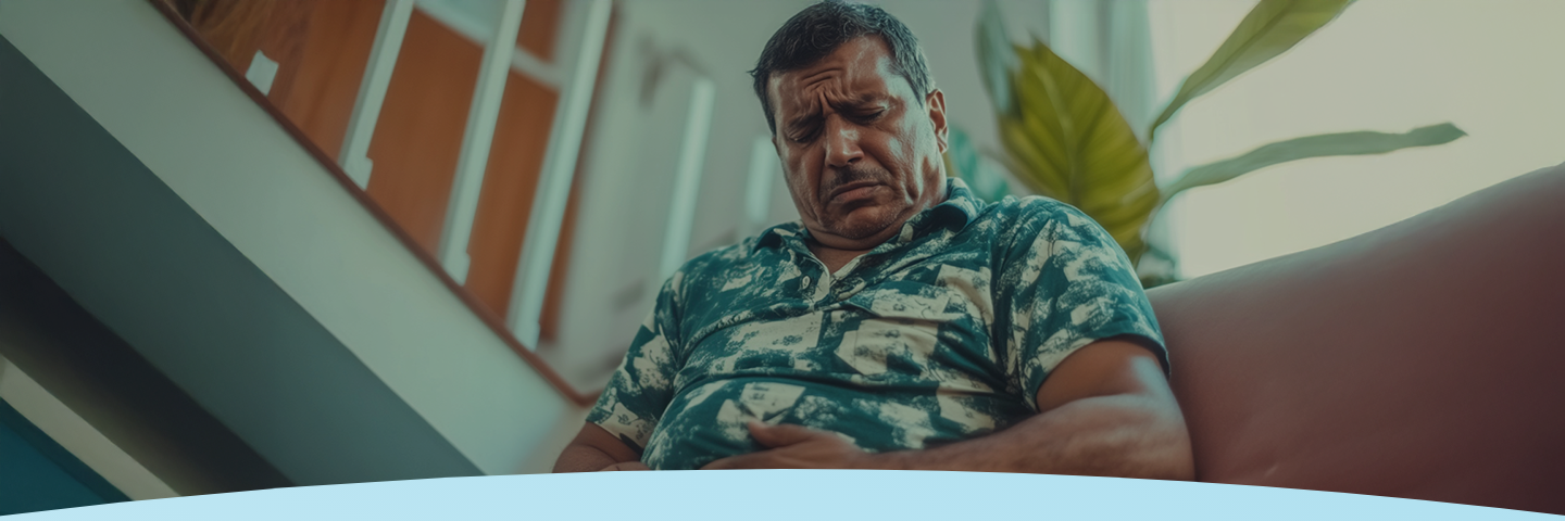 Homem sentado em um sofá segurando o estômago com uma expressão de dor.