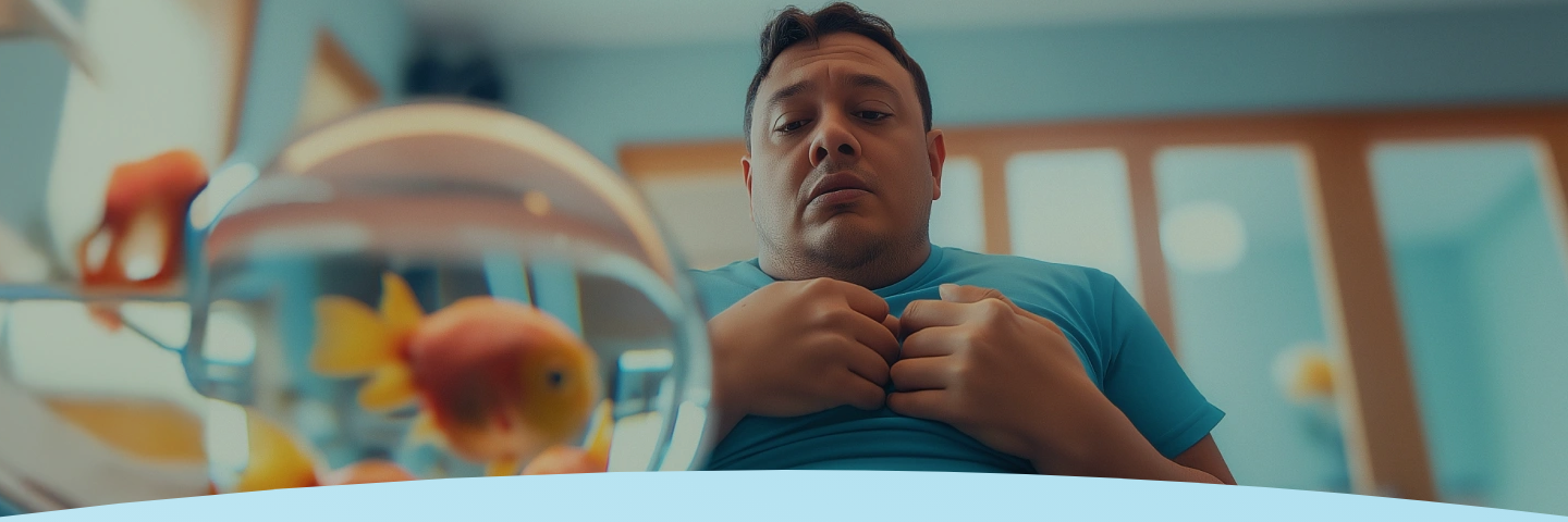 Homem de camisa azul segura o peito, sentado perto de um aquário com peixes dourados.