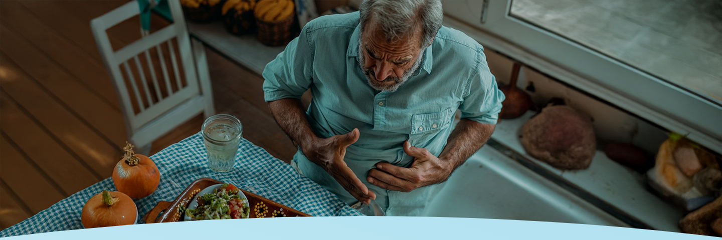 Homem idoso sentado em uma mesa, segurando o estômago com desconforto.