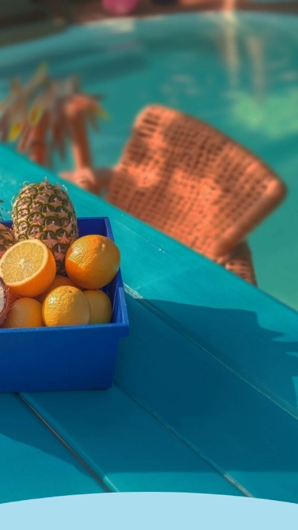 Frutas tropicais em uma caixa azul em uma mesa turquesa ao lado de uma piscina com uma bola rosa.